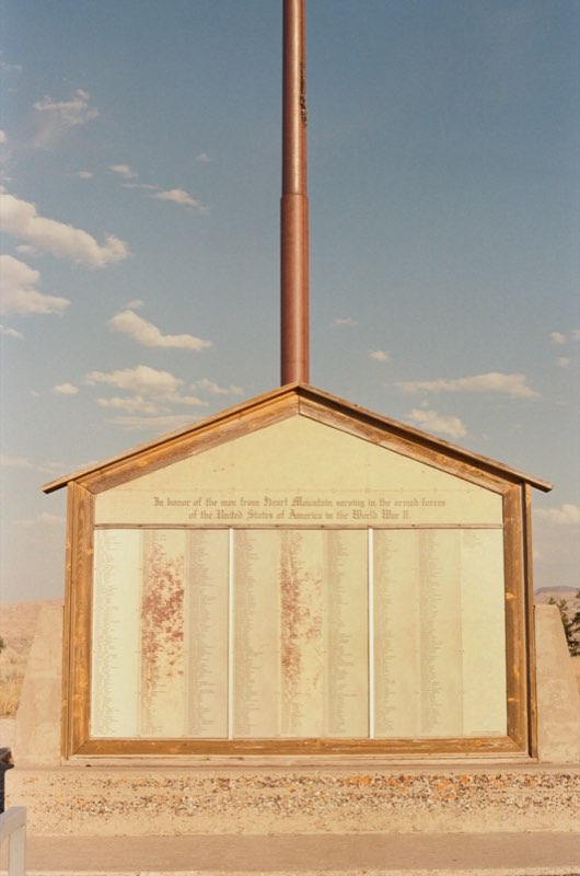A weathered wooden honor roll under a gabled roof and a tall flagpole, listing the men from Heart Mountain who served in World War II.