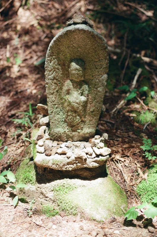 A mossed stone stele carved with a standing figure, its plinth heaped with small stones, pine cones and coins.
