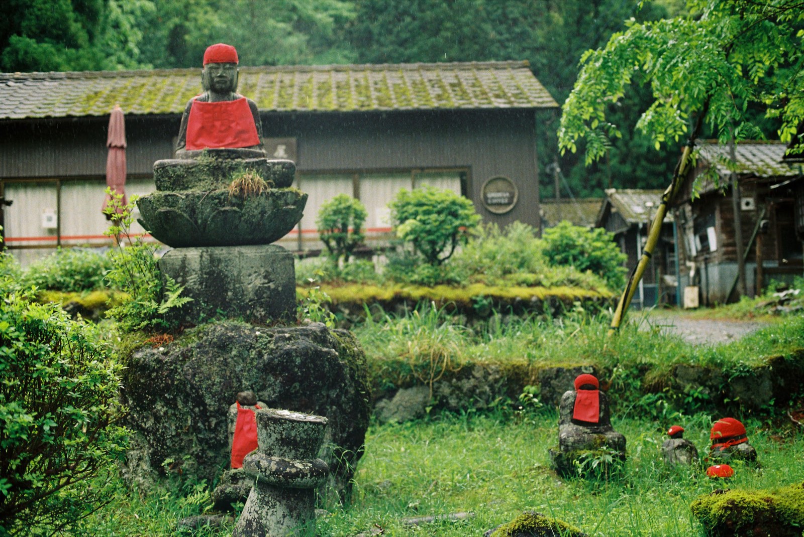 Stone figures in red bibs and caps standing in wet grass in the rain, a moss-tiled village roof behind them.