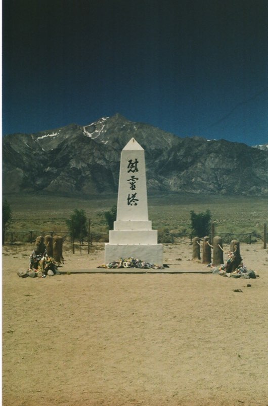 A white obelisk lettered in Japanese stands in the desert below snow-streaked mountains, strings of paper cranes heaped at its base and on the posts to either side.
