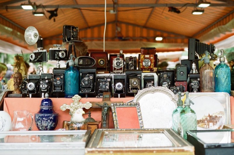 A market table crowded with old cameras, crucifixes and glass siphons.