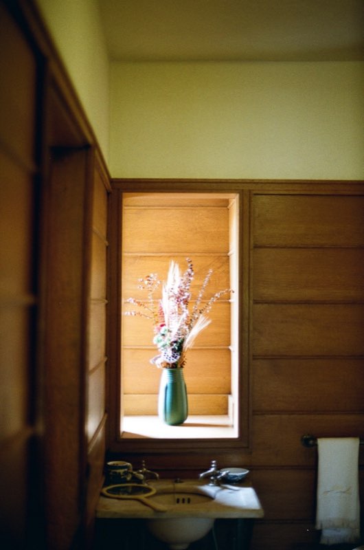 A green vase of dried grasses and flowers set in a sunlit niche in an oak-paneled wall, above an old washstand.