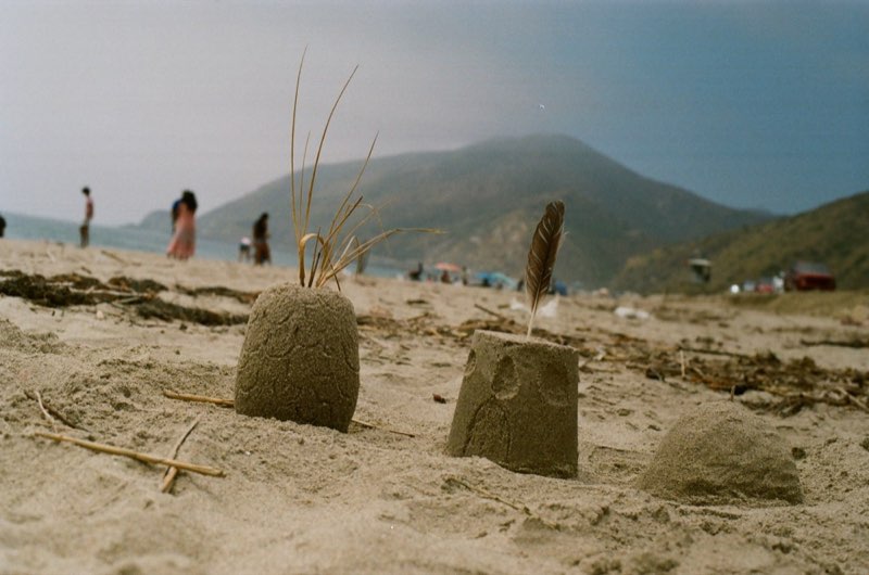 Three small sand figures on a beach, one crowned with dry grass and one with a feather, people and a headland blurred behind.