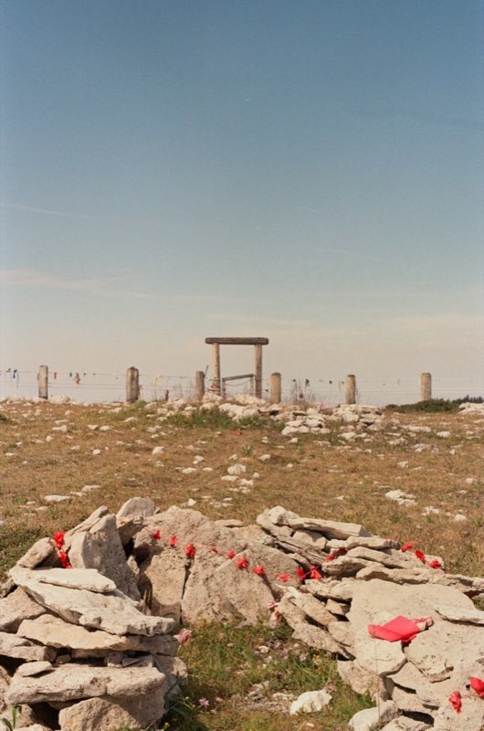 A cairn of flat stones strung with red rags, a wooden gate on the ridge behind.